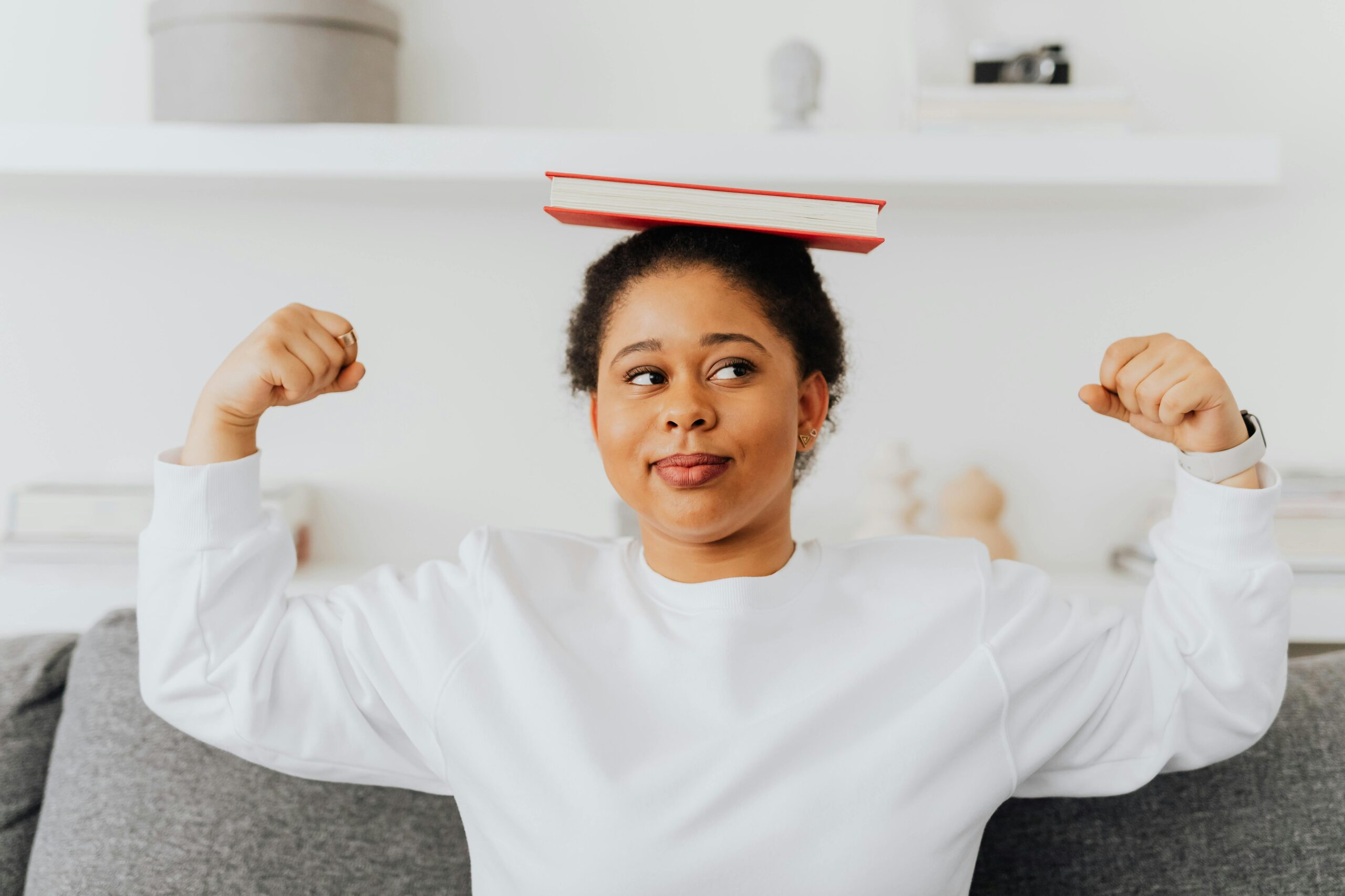 A confident woman balances a book on her head and flexes indoors. Casual and relaxed atmosphere.